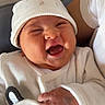 baby, smiling, infant, white_clothing, hat, hand, person, indoor, cute, joyful, newborn, face, child, holding, happy, cozy, human, apparel, portrait, sitting