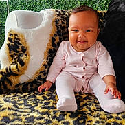 Journey is registered to the contest to win money with this photo: baby, child, smiling, pink_clothing, onesie, leopard_print, blanket, chair, plush, fur, greenery, plants, indoor, portrait, happy, sitting, face, cute, infant, cozy