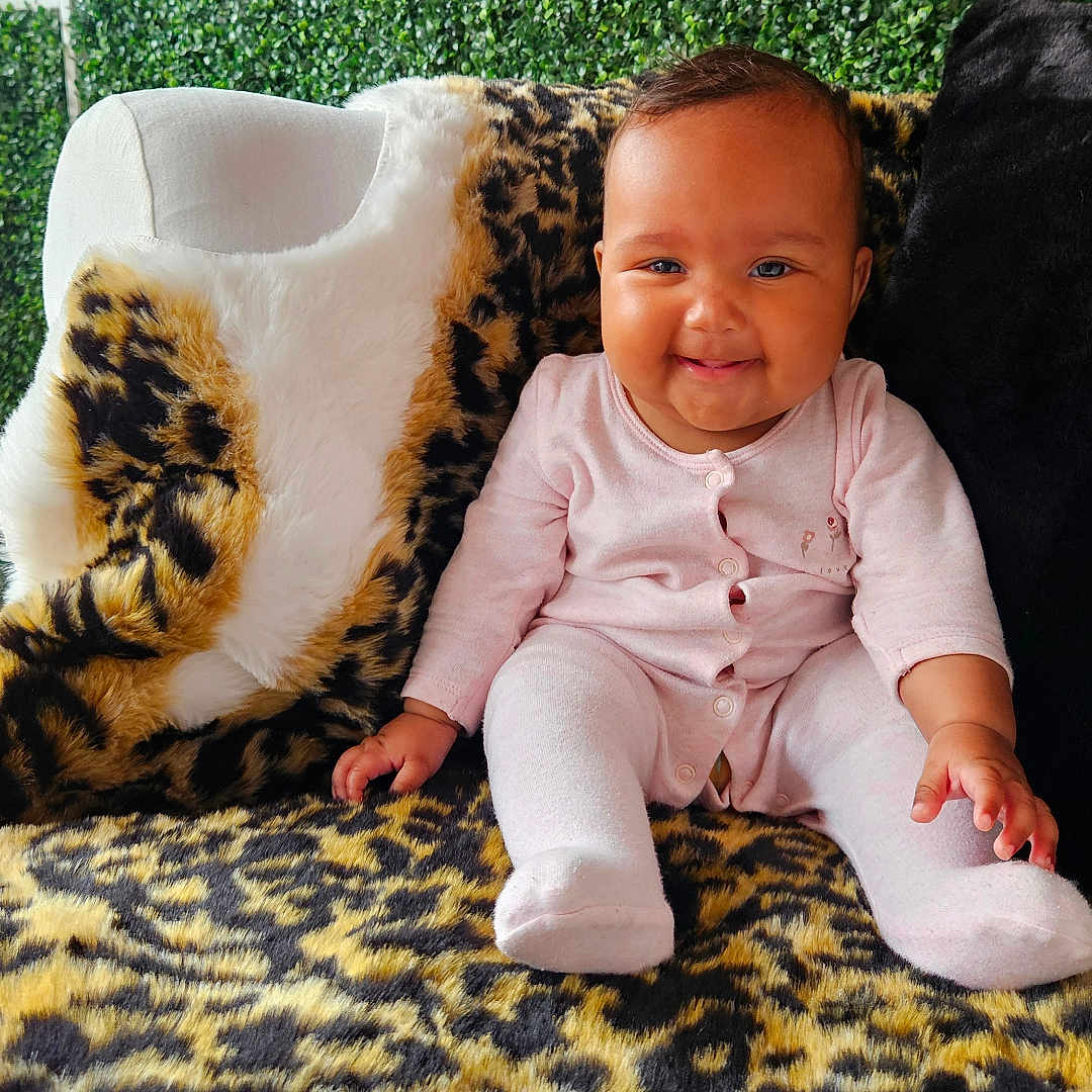 Journey is registered to the contest to win money with this photo: baby, blanket, chair, child, cozy, cute, face, fur, greenery, happy, indoor, infant, leopard_print, onesie, pink_clothing, plants, plush, portrait, sitting, smiling