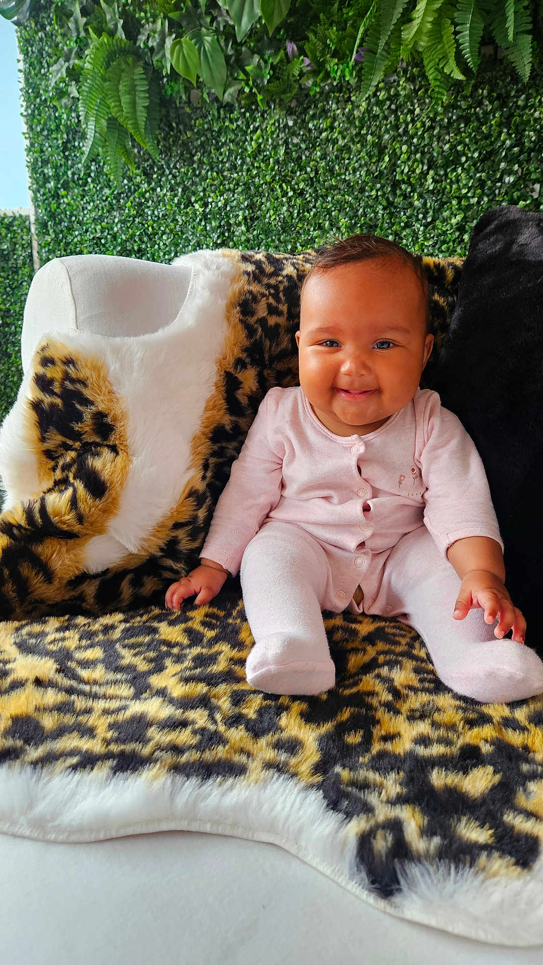 Journey is registered to the contest to win money with this photo: baby, child, smiling, pink_clothing, onesie, leopard_print, blanket, chair, plush, fur, greenery, plants, indoor, portrait, happy, sitting, face, cute, infant, cozy