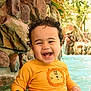 toddler, child, smiling, water, pool, yellow_shirt, curly_hair, happy, outdoor, rocks, greenery, sitting, splashing, sunlight, swimwear, cute, young_child, water_fun, portrait, playful