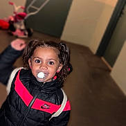Zeyna a rejoint le concours — aidez-le/la à gagner de superbes lots ! black_jacket, child, clothing, curly_hair, cute, door, earrings, floor, indoor, nike_logo, pacifier, pants, person, pink_stripes, plush_toy, portrait, smiling, toddler, tricycle, young_child