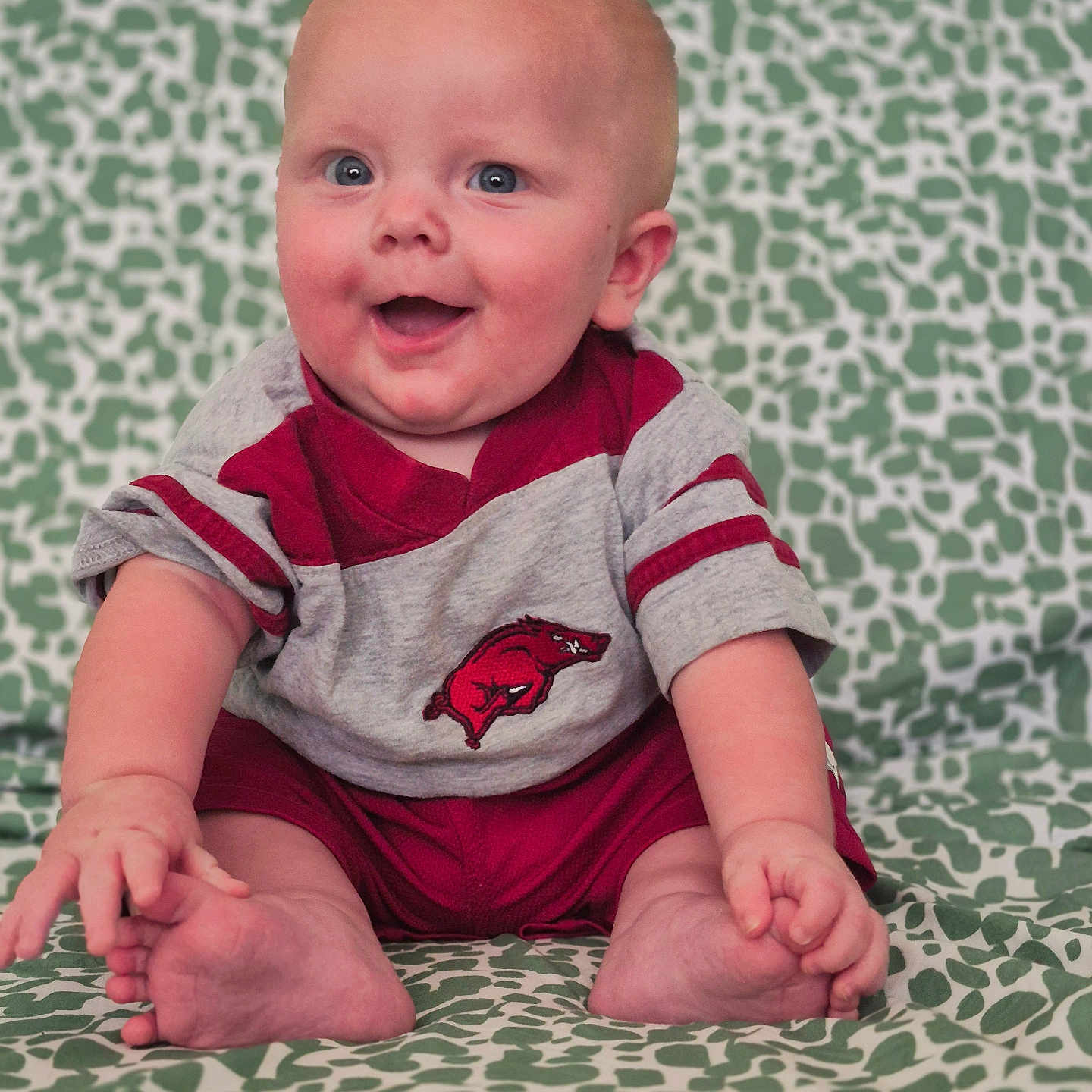 Opie is registered to the contest to win money with this photo: baby, blue_eyes, child, clothing, cute, face, foot, gray_clothing, hand, happy, indoor, infant, patterned_blanket, person, portrait, red_clothing, sitting, skin, smiling, young_child