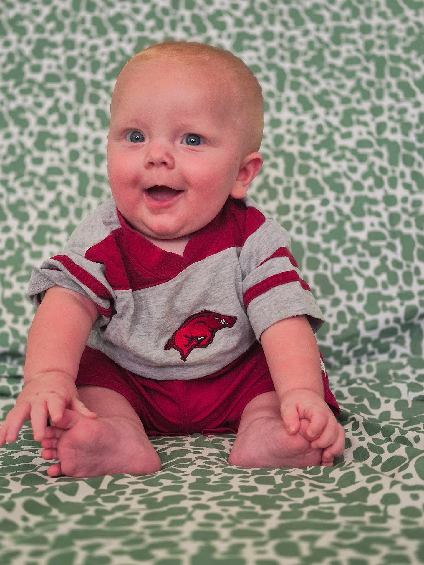 Opie is registered to the contest to win money with this photo: baby, child, smiling, sitting, clothing, patterned_blanket, happy, infant, cute, portrait, hand, foot, indoor, person, young_child, red_clothing, gray_clothing, skin, face, blue_eyes