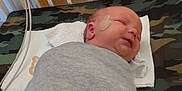 Walker is registered to the contest to win money with this photo: newborn, baby, swaddled, sleeping, crib, hospital, medical_patch, blanket, towel, camouflage, infant, bed, wooden_bars, resting, cute, small, face, person, indoors, healthcare