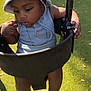 baby, black_swing, bucket_hat, chain, child, cute, fence, footwear, gold_chain, green_turf, hat, leaves, outdoor, person, playground, sandal, summer_clothing, sunlight, swing, toddler