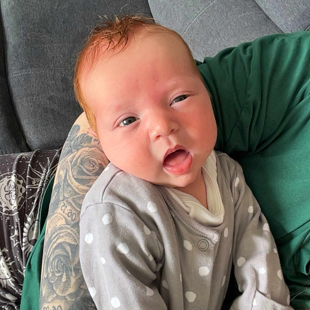 Brodie is registered to the contest to win money with this photo: arm, baby, casual, comfort, cute, gray_couch, green_shirt, holding, indoor, infant, open_mouth, patterned_pants, person, polka_dot_clothing, red_hair, relaxing, seated, skin, tattoo, young_child