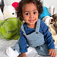 Landon is registered to the contest to win money with this photo: toddler, child, curly_hair, blue_clothing, overalls, white_socks, bed, plush_toys, green_turtle, stuffed_animal, smiling, indoors, cute, portrait, person, happy, soft_toys, childhood, playful, cozy
