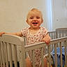 Oaklynn-Ayla is registered to the contest to win money with this photo: toddler, child, crib, baby, smile, standing, onesie, indoor, toy, room, furniture, happy, blonde_hair, baby_clothing, playroom, baby_bed, wall_shelf, decor, infant, person