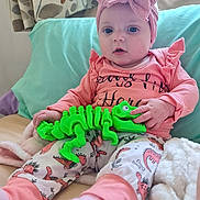 Oaklynn-Ayla joined the competition — help win amazing prizes! baby, child, pink_clothing, headband, bow, toy, green_toy, dinosaur_toy, sitting, blanket, pillow, bed, indoors, cute, infant, soft_toys, playful, cozy, person, portrait