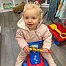 bells, bookshelf, child, curly_hair, floor, flooring, happy, indoor, person, pink_clothing, playroom, playtime, ride_on_vehicle, shovel, smiling, stuffed_toy, toddler, toy, toy_shelf, wagon