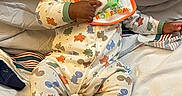 Jelani is registered to the contest to win money with this photo: baby, sunglasses, bib, onesie, dinosaur_pattern, bed, blanket, pillow, infant, clothing, child, person, cute, cozy, indoor, sleepwear, footed_pajamas, playful, resting, toddler