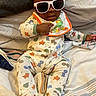 baby, sunglasses, bib, onesie, dinosaur_pattern, bed, blanket, pillow, infant, clothing, child, person, cute, cozy, indoor, sleepwear, footed_pajamas, playful, resting, toddler