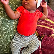 Jelani is registered to the contest to win money with this photo: baby, sleeping, infant, red_onesie, striped_pants, pizza_hat, red_socks, pillow, blanket, indoor, cute, resting, child, peaceful, cozy, soft_fabric, headwear, lying_down, small_hands, footwear