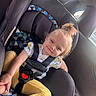 child, toddler, car_seat, car_interior, seatbelt, smile, happy, yellow_pants, floral_shirt, hair_tie, window, sunlight, safety, baby, cute, face, person, seat, restraint, travel