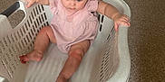 Willow is registered to the contest to win money with this photo: baby, child, laundry_basket, pink_clothing, carpet, indoor, floor, furniture, playmat, hand, foot, curious, sitting, home, person, infant, looking_down, grip, white, plastic