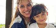 Ezra is registered to the contest to win money with this photo: woman, child, smile, embrace, outdoor, sports_jersey, number_19, white_tshirt, long_hair, curly_hair, portrait, happy, people, casual_clothing, daylight, roof, pole, background, parking_lot, vehicle