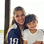 woman, child, smile, embrace, outdoor, sports_jersey, number_19, white_tshirt, long_hair, curly_hair, portrait, happy, people, casual_clothing, daylight, roof, pole, background, parking_lot, vehicle
