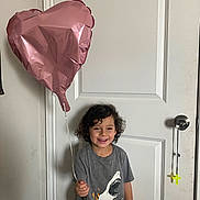 Ezra joined the competition — help win amazing prizes! child, boy, smiling, indoor, balloon, heart_shaped, curly_hair, grey_shirt, dinosaur_graphic, door, doorknob, casual_clothing, happy, standing, young, person, toy, string, wall, light