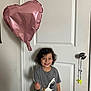 child, boy, smiling, indoor, balloon, heart_shaped, curly_hair, grey_shirt, dinosaur_graphic, door, doorknob, casual_clothing, happy, standing, young, person, toy, string, wall, light