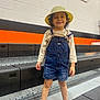 Ezra joined the competition — help win amazing prizes! child, barefoot, denim_overalls, yellow_hat, smiling, gymnasium, bleachers, orange_wall, black_seats, curly_hair, standing, casual_clothing, indoor, happy, person, young_child, playful, bright_colors, portrait, full_body