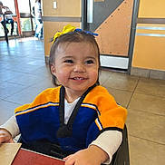 Arianna joined the competition — help win amazing prizes! child, toddler, smiling, happy, indoor, restaurant, table, clothing, hair_bow, yellow, blue, sweater, face, person, seat, window, tile_floor, door, light, casual