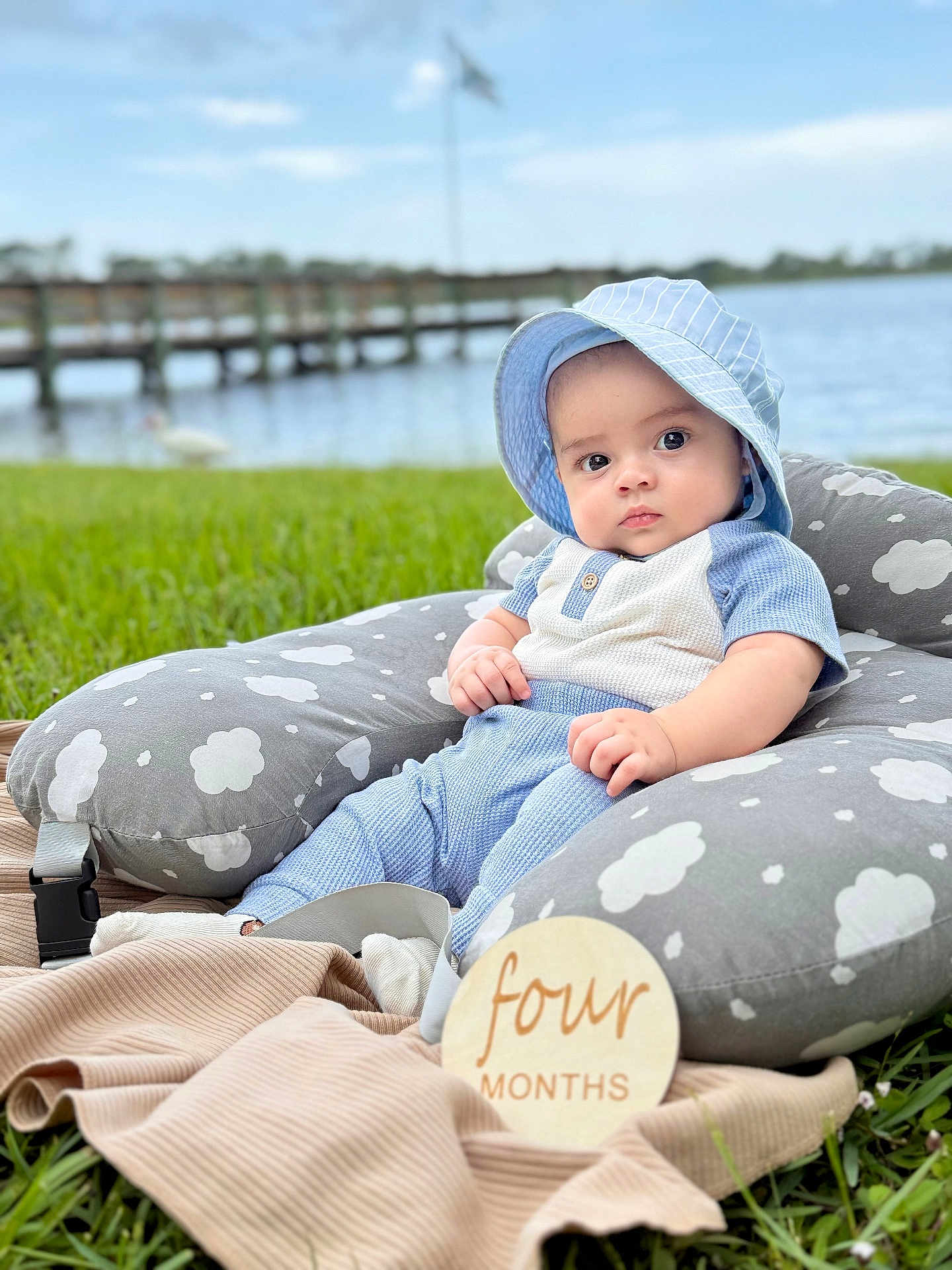 Zayn Loaiza is registered to the contest to win money with this photo: baby, infant, blue_clothing, sunhat, pillow, cloud_pattern, grass, outdoor, lake, pier, windmill, wooden_sign, four_months, milestone, cute, portrait, child, sitting, blanket, nature