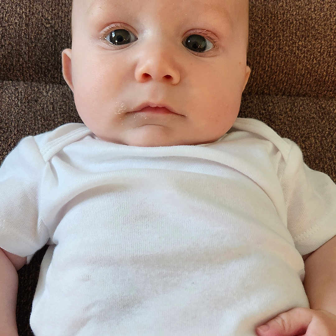 Ethan is registered to the contest to win money with this photo: baby, bodypart, clothing, couch, cushion, face, finger, frown, furniture, hand, head, homedecor, newborn, pants, person, photography, portrait, sad, sitting, t_shirt