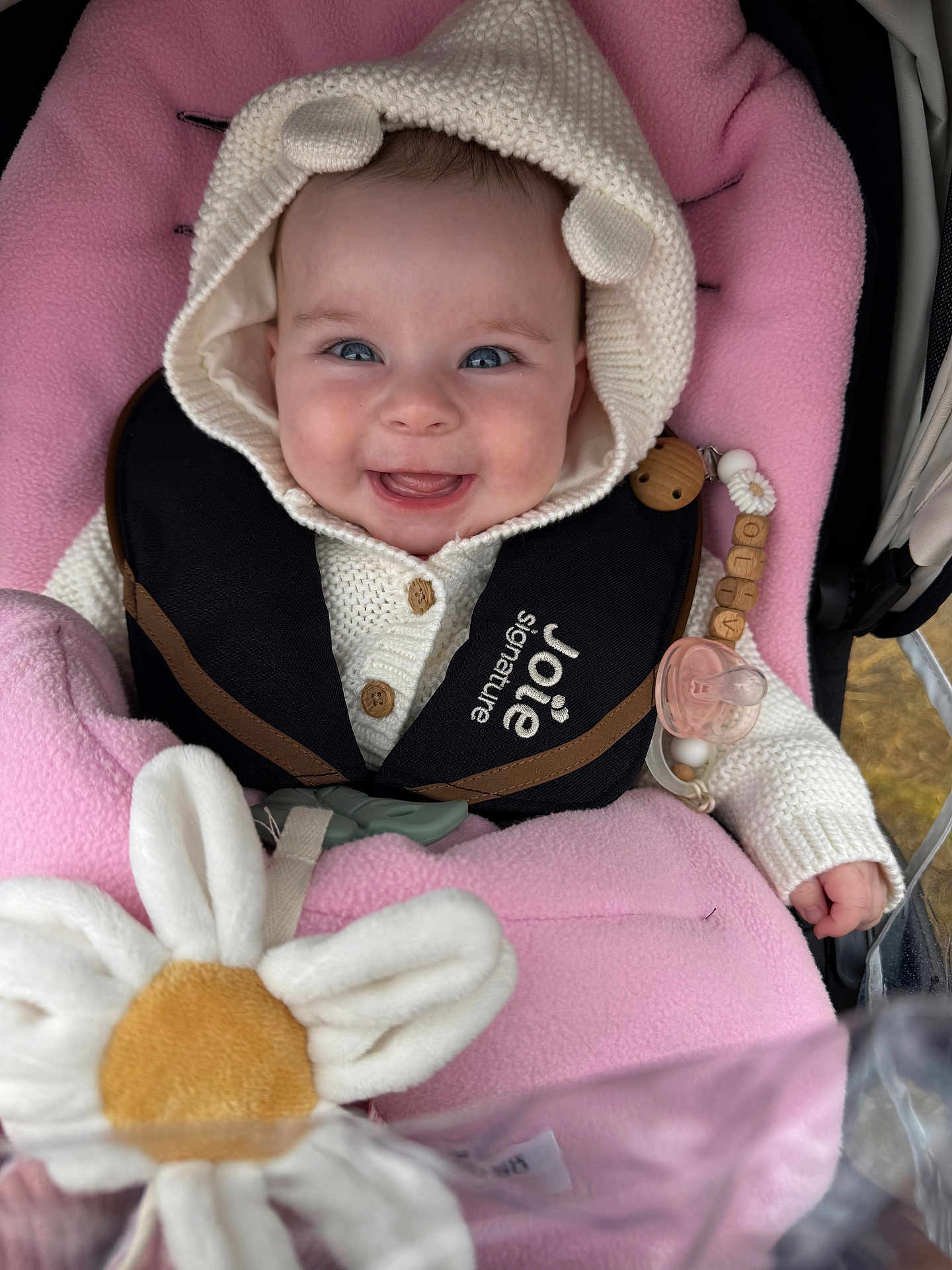 Olivia is registered to the contest to win money with this photo: baby, smiling, knitted_hoodie, bear_ears, pink_blanket, stroller, plush_toy, pacifier, baby_clothing, child, happy, cute, infant, seat_harness, wooden_beads, toy_flower, closeup, face, portrait, baby_accessories