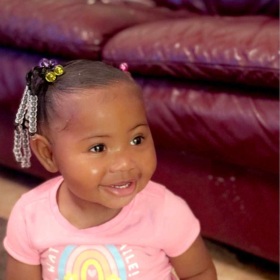 Ja'Ryiah is registered to the contest to win money with this photo: bright_eyes, child, clothing, couch, cute, face, hair_beads, happy, indoor, leather_furniture, person, pink_shirt, portrait, rainbow_graphic, short_hair, sitting, skin, smiling, toddler, young_child