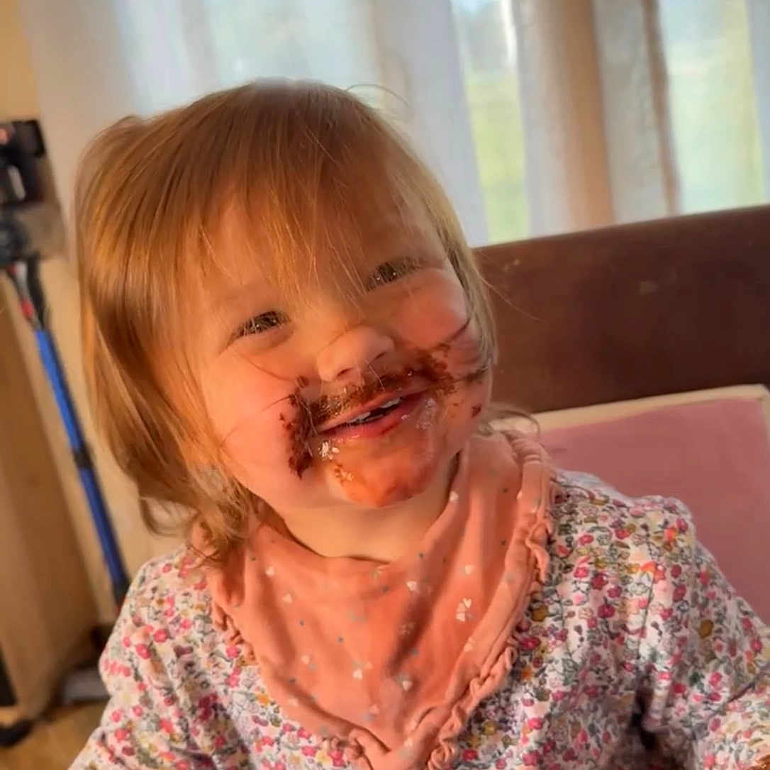 émiliana a rejoint le concours — aidez-le/la à gagner de superbes lots ! bib, cheeky, child, chocolate, curtains, cute, face, floral_clothing, food, hair, happy, indoor, messy_face, playful, sitting, smiling, sunlight, table, toddler, young_child