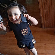 Amy is registered to the contest to win money with this photo: toddler, child, indoor, barefoot, phone, leopard_print, black_clothing, smile, wooden_floor, rug, hair, person, home, cute, holding, standing, furniture, room, happy, young_child