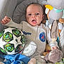 baby, infant, plush_toy, soccer_ball, onesie, bassinet, pacifier, soft_toys, child, cute, surprised_expression, person, indoor, baby_clothes, stuffed_animal, toy, smiling, small_child, sleeping_area, comfort