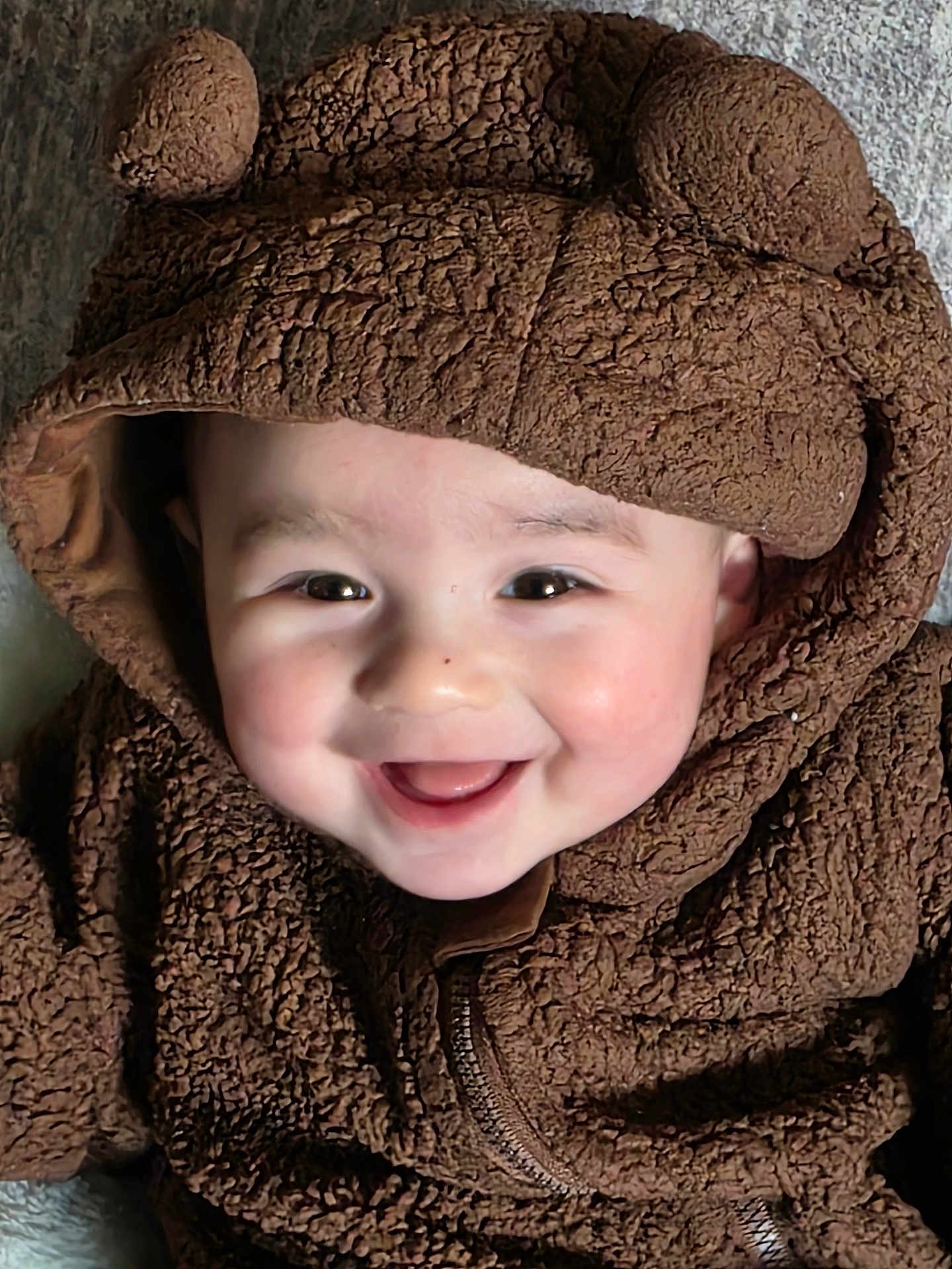Jack is registered to the contest to win money with this photo: baby, infant, child, smiling, hood, brown_fur, fuzzy_jacket, cute, rosy_cheeks, close_up, portrait, eyes, teeth, happy, cozy, zipper, fabric_texture, warm_clothing, blanket, laying_down
