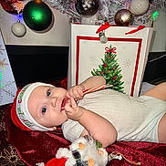 Carson joined the competition — help win amazing prizes! baby, infant, child, candy_cane, santa_hat, christmas_tree, ornament, gift_bag, plush_toy, holiday, festive, lying_down, blanket, onesie, blue_eyes, red_decor, tree_skirt, decorations, face, person