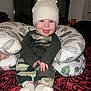 Carson joined the competition — help win amazing prizes! baby, infant, toddler, knit_hat, hat, smile, blue_eyes, pillow, patterned_pillow, camouflage_clothing, camo_pants, onesie, socks, bedspread, blanket, indoor, portrait, sitting, hands, feet