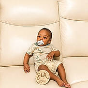Josiah joined the competition — help win amazing prizes! baby, infant, pacifier, couch, leather, milestone, six_months, plaque, clothing, dinosaur_print, child, person, sitting, indoors, cute, young, skin, foot, hand, relaxing