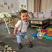 Mayron a rejoint le concours — aidez-le/la à gagner de superbes lots ! toddler, child, smiling, playful, indoor, living_room, toy, colorful, suspenders, bow_tie, shorts, sandals, couch, pillows, curtains, table, high_chair, floor, tiles, home