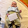 Léo a rejoint le concours — aidez-le/la à gagner de superbes lots ! baby, infant, smiling, blanket, bib, sneakers, shoes, clothing, cute, face, person, floor, sign, handwritten, indoor, soft_texture, orange, white, green, young_child