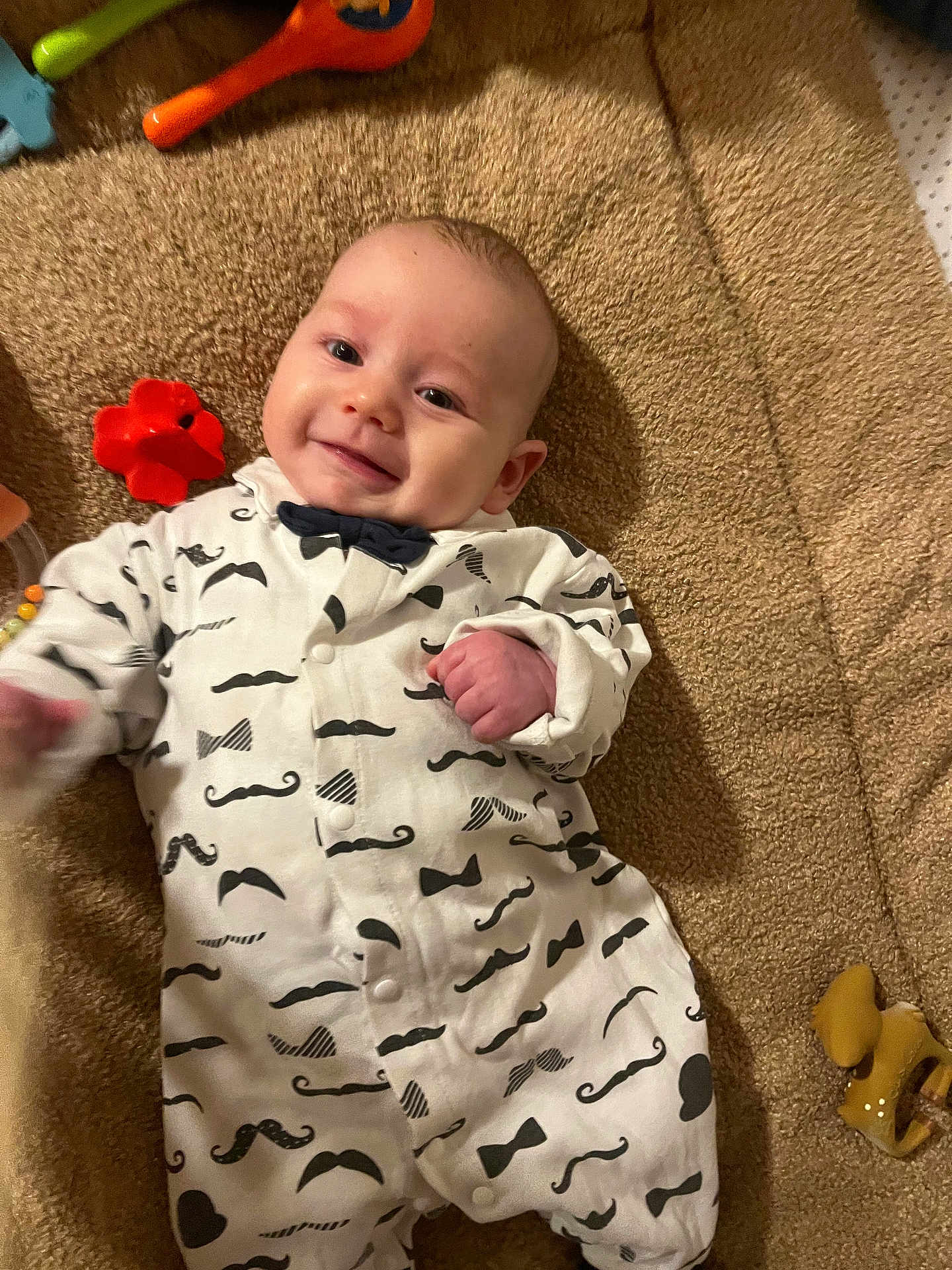 Léo a rejoint le concours — aidez-le/la à gagner de superbes lots ! baby, infant, smiling, onesie, mustache_pattern, bow_tie_pattern, carpet, toy, red_toy, yellow_toy, soft_texture, cute, child, happy, lying_down, indoor, playtime, clothing, face, person