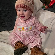 Rachel is registered to the contest to win money with this photo: baby, blanket, boots, brown_boots, child, cozy, cute, happy, hat, home, indoors, infant, knitted_hat, pink_clothing, pom_pom, portrait, sitting, smiling, warm_clothing, white_blanket