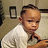 toddler, child, cornrows, white_shirt, table, game_controller, indoor, carpet, curious, leaning, person, young_child, casual_clothing, home, portrait, brown_skin, short_sleeves, looking_away, hand, expression