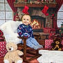 blanket, blue_sweater, child, christmas_decor, curtains, festive, fireplace, holiday, indoor, jeans, plush_toy, smiling, socks, stockings, sweater, teddy_bear, toddler, wooden_chair, wrapped_gifts, wreath