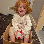 Andie joined the competition — help win amazing prizes! child, red_hair, curly_hair, smiling, cardboard_box, soccer_ball, indoor, floor, door, white_clothing, long_sleeve, pants, happy, playful, person, toy, young_child, casual, home, sitting