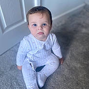 Vinny is registered to the contest to win money with this photo: baby, child, pajamas, carpet, floor, indoors, blue_eyes, sitting, white_clothing, curly_hair, door, soft_light, portrait, infant, cute, face, person, young_child, home, expression