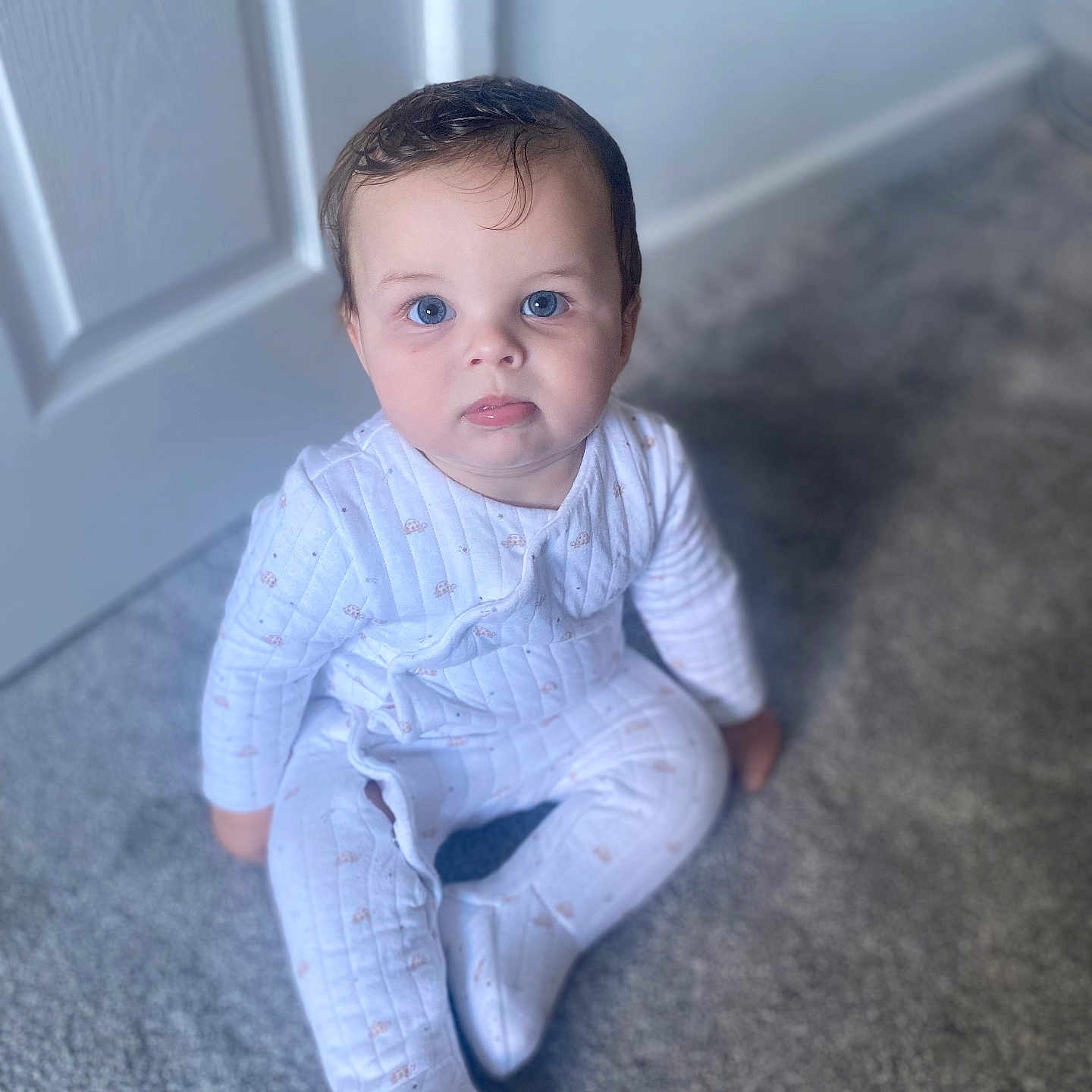 Vinny is registered to the contest to win money with this photo: baby, blue_eyes, carpet, child, curly_hair, cute, door, expression, face, floor, home, indoors, infant, pajamas, person, portrait, sitting, soft_light, white_clothing, young_child