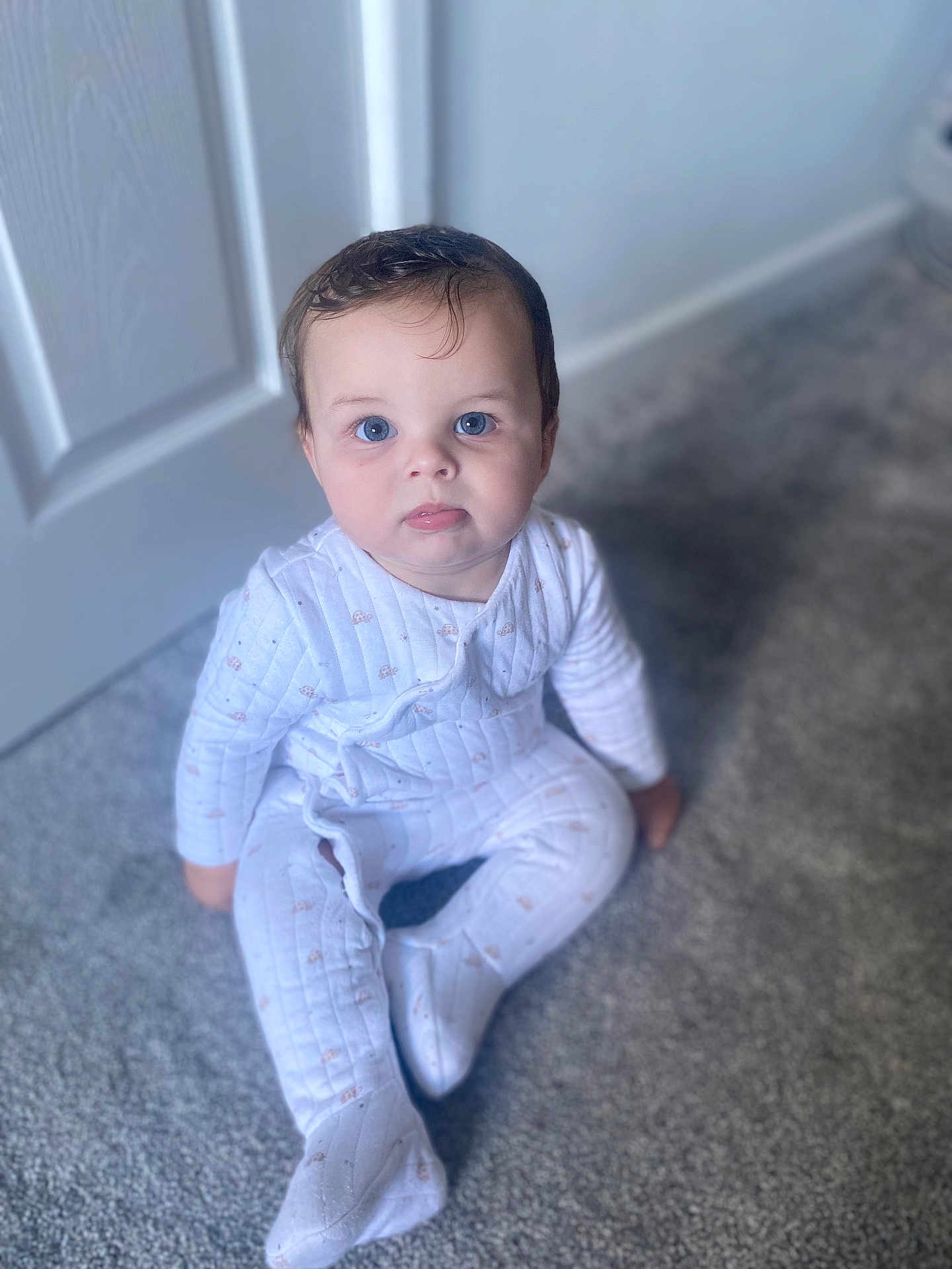 Vinny is registered to the contest to win money with this photo: baby, child, pajamas, carpet, floor, indoors, blue_eyes, sitting, white_clothing, curly_hair, door, soft_light, portrait, infant, cute, face, person, young_child, home, expression