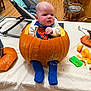 baby, pumpkin, superman_outfit, table, pumpkin_carving, blue_socks, indoor, wood_floor, chair, spoon, green_scraper, orange_pumpkin, baby_face, costume, festive, halloween, child, cute, carving_tools, person