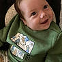 baby, child, smiling, green_sweater, dinosaur_print, pants, cushioned_seat, face, eyes, infant, clothing, cute, happy, portrait, young_child, sitting, indoors, apparel, baby_fashion, closeup