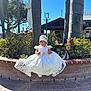 toddler, child, white_dress, lace, bonnet, smile, brick_wall, garden, plants, statue, tree, palm_tree, outdoor, sunny, blue_sky, person, flower, shoes, portrait, daylight